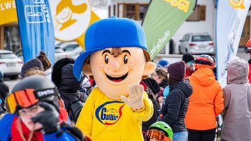 A cheerful mascot wearing a blue hat and yellow sweater stands in the midst of a group of people on a ski resort. In the background, colorful flags and snow-covered mountains can be seen.