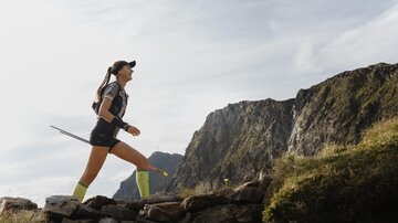 A female runner jogging on a mountain path, surrounded by impressive rocks and a wide view. She is wearing athletic clothes and has a walking stick in her hand.