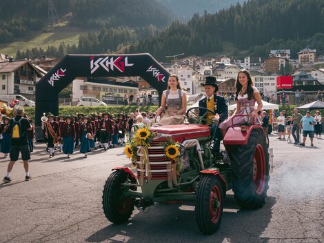 A lively event featuring a tractor accompanied by two women in traditional costumes. In the background, cheering people and a picturesque mountain landscape can be seen.