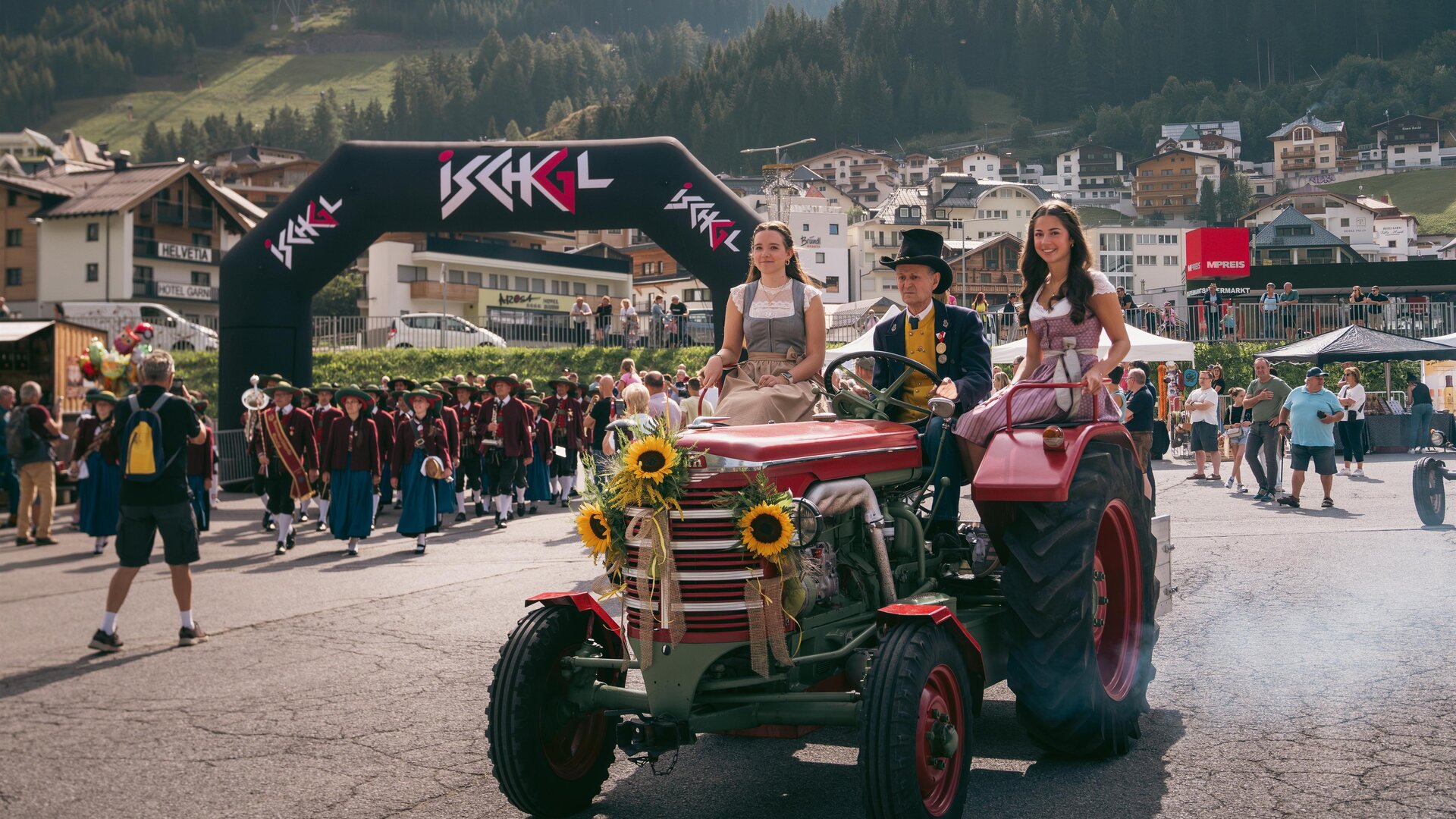 A lively event featuring a tractor accompanied by two women in traditional costumes. In the background, cheering people and a picturesque mountain landscape can be seen.
