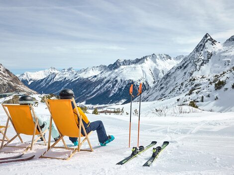 A cozy ski resort with sun loungers and snow-covered mountains in the background. Two people relax in the loungers while enjoying the view.