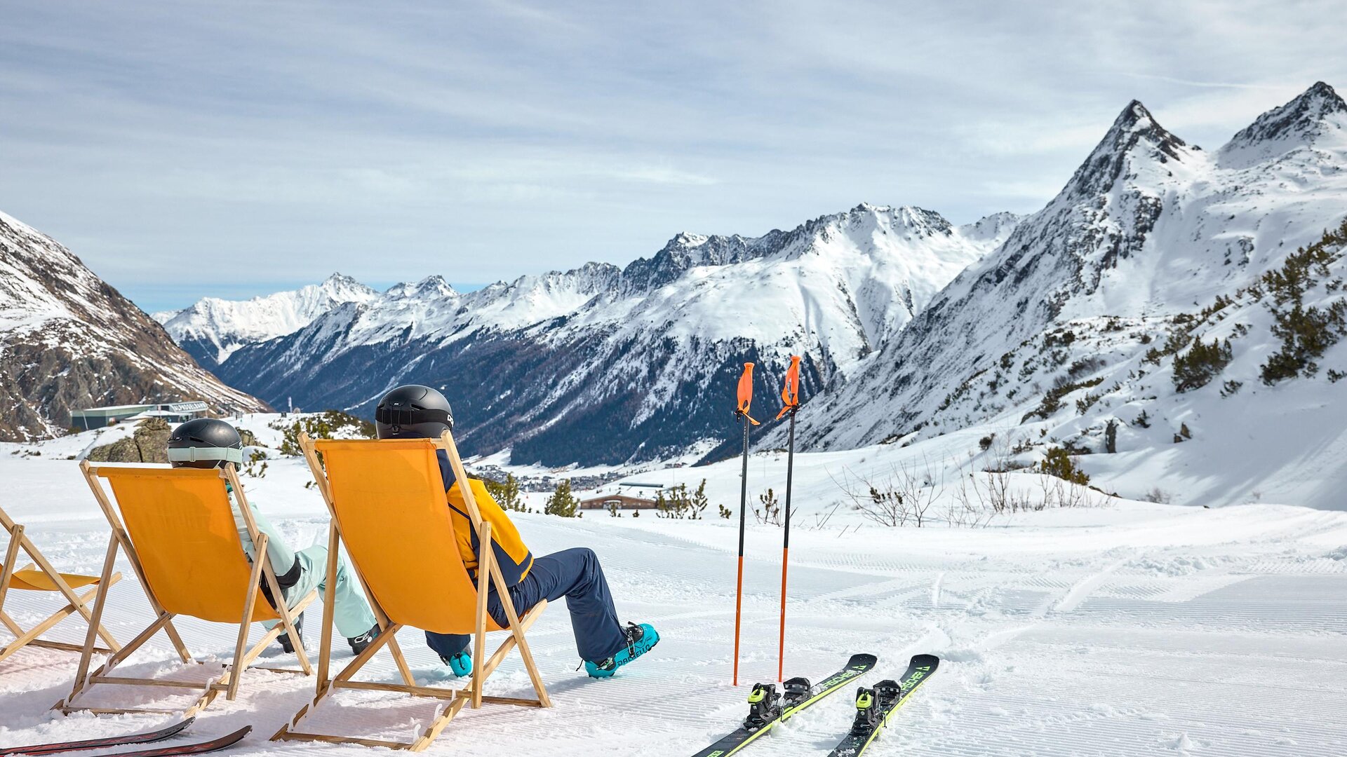 A cozy ski resort with sun loungers and snow-covered mountains in the background. Two people relax in the loungers while enjoying the view.