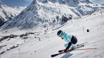 A skier descends the slope, surrounded by snow-covered mountains. The sunny scene features clear blue skies and a vibrant winter landscape.