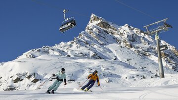 Two skiers are skiing on a snow-covered slope under a clear blue sky. In the background, there is a mountain and a gondola.