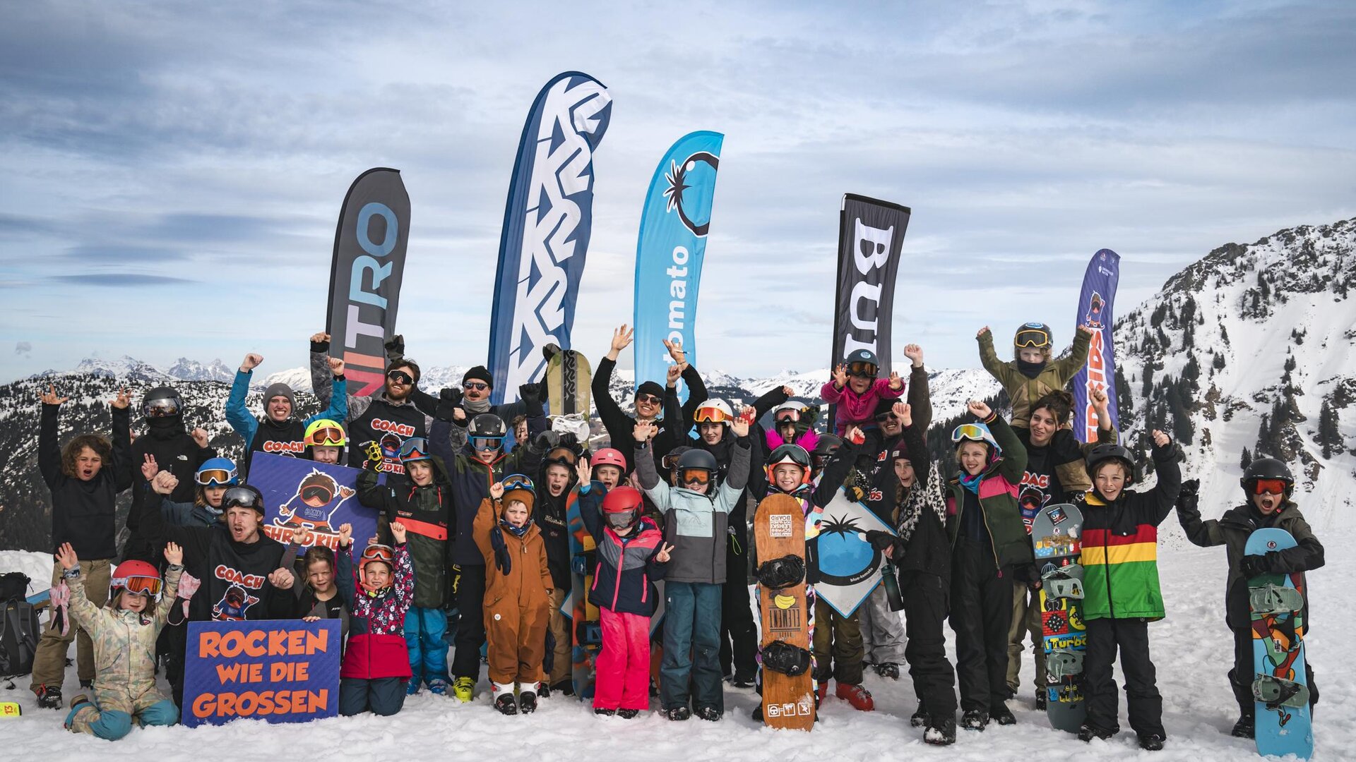 A group of children and adults stands happily in the snow. They are wearing ski equipment and are celebrating with flags in the background.