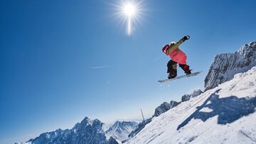 A snowboarder jumps over a snow-covered slope. The sky is blue and the sun shines brightly.