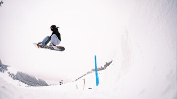 A snowboarder jumps high in the air over a snowy slope. In the background, trees and a wintry landscape can be seen.