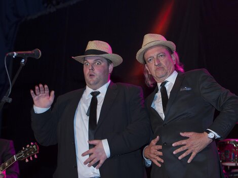 Two men in suits and hats are posing amusingly on stage. In the background, musicians with instruments can be seen.