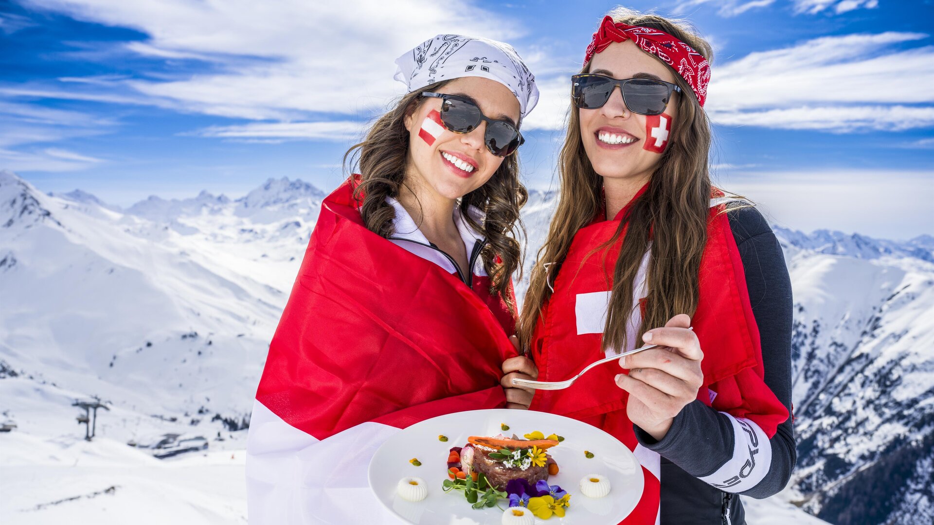 Two women in sunglasses are standing on a snow-covered mountain, holding a tray of food. They are wearing flags of Switzerland and smiling in front of an impressive mountain landscape.
