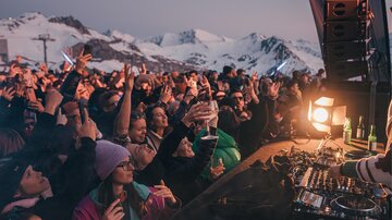 An enthusiastic crowd dances at a festival in the mountains. In the background, snow-covered peaks and an atmospheric sunset can be seen.