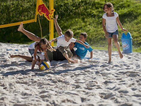 A group of children is happily playing volleyball in the sand. Two children are lying in the sand while the others are playing around the ball.