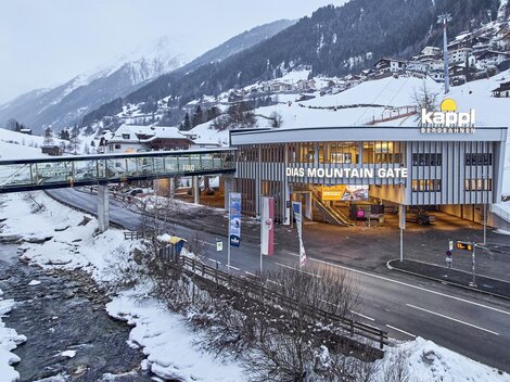 Ein modernes Gebäude mit der Aufschrift "DAS MOUNTAIN GATE" steht in einer schneebedeckten Landschaft. Im Hintergrund sind Berge und ein Fluss zu sehen.