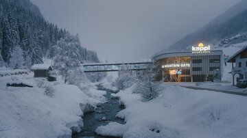 A snowy winter landscape with a river and trees. In the background, there is a modern building with a glowing sign.
