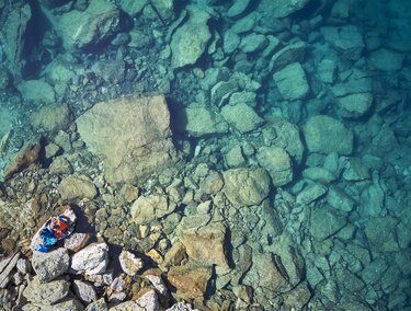 A clear blue lake with many large stones at the bottom. A person is lying relaxed on the shore.