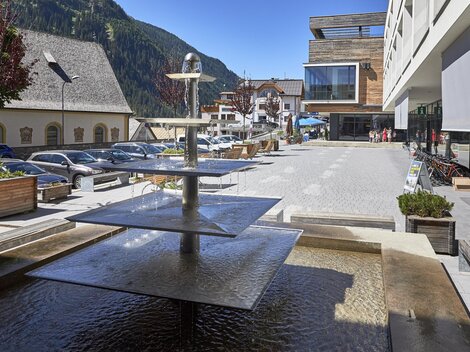 A modern square with a water basin and several levels. In the background, cars and buildings can be seen, surrounded by mountains.