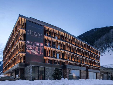 A modern hotel with a stylish facade and illuminated balconies. Surrounded by snow-covered mountains and a clear evening sky.
