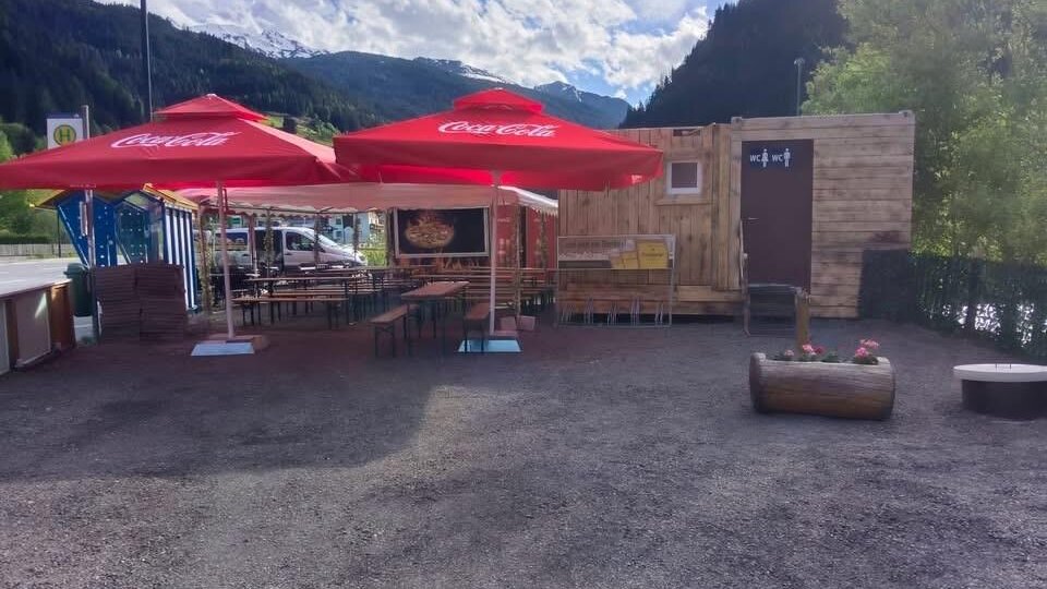 A small snack stand with a wooden structure and red umbrellas. The surroundings are mountainous and green, ideal for a break outdoors.
