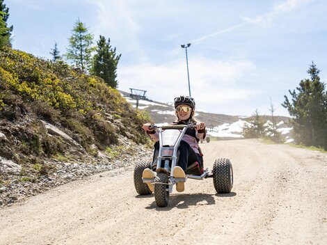 Ein Kind fährt mit einem Fahrzeug auf einer unbefestigten Straße in den Bergen. Im Hintergrund sind Bäume und eine sonnige Landschaft zu sehen.