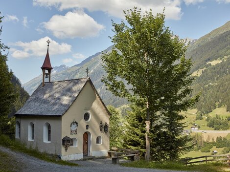 A small chapel surrounded by trees and mountains. The sky is partly cloudy and the landscape is green and picturesque.