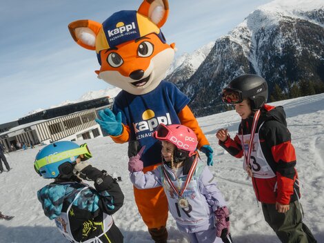 Ein Maskottchen in einem blauen Shirt und einer Mütze steht auf einem schneebedeckten Hang. Um es herum stehen drei Kinder in Skiausrüstung und feiern zusammen.