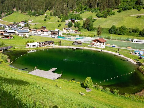 Ein malerischer Teich umgeben von grünen Wiesen und Bergen. Im Hintergrund sind mehrere Häuser und Anlagen sichtbar.